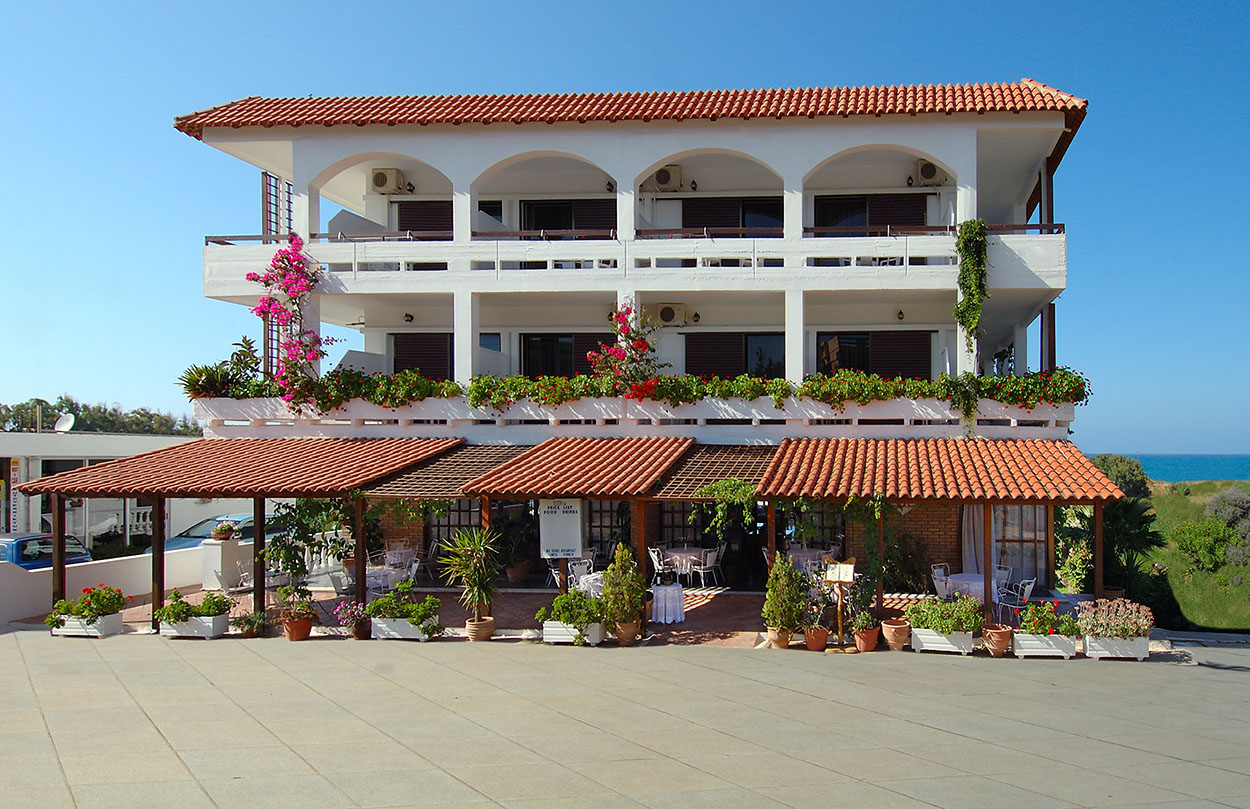 
White three-story hotel building with terracotta roof, balconies and flowering plants in Maleme