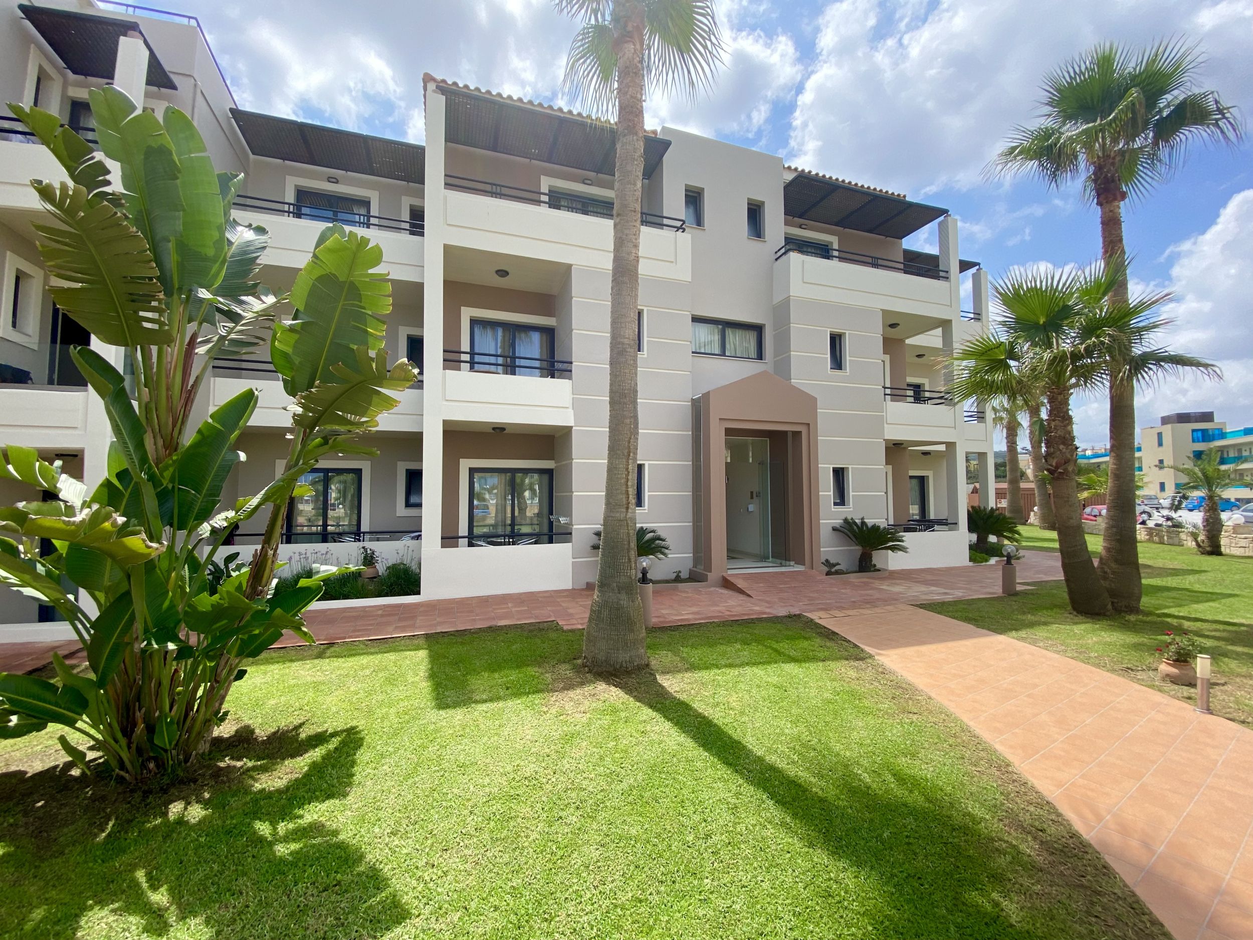 
Three-story apartment building with balconies, palm trees and green lawn under a bright sky