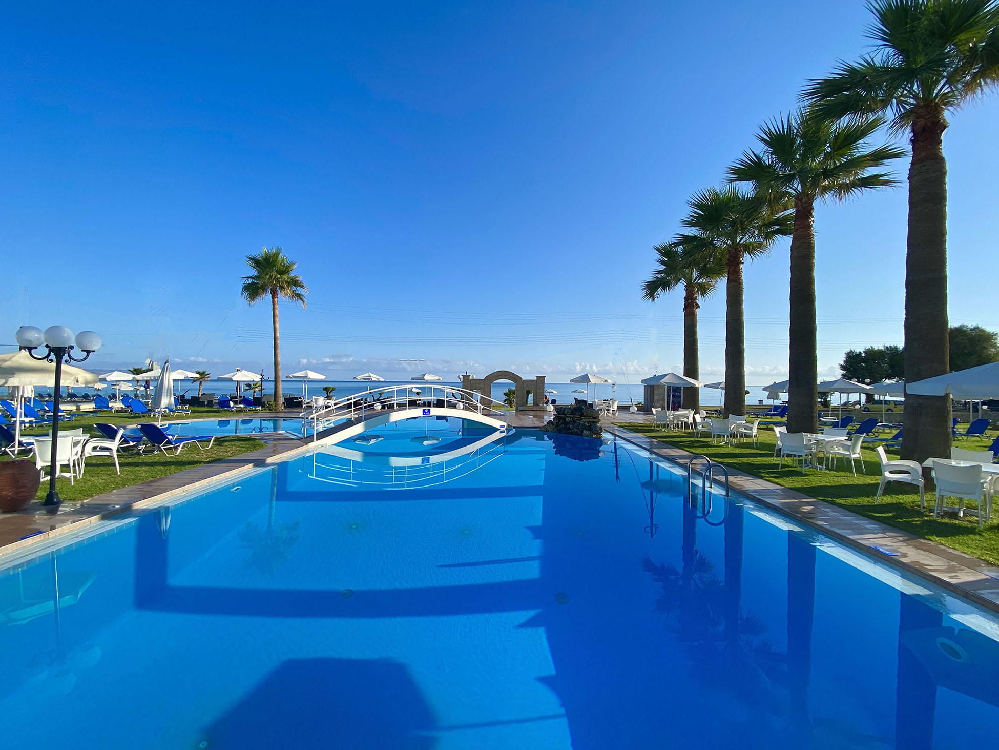 
Large outdoor swimming pool with palm trees, sunbeds and a small white bridge under blue sky