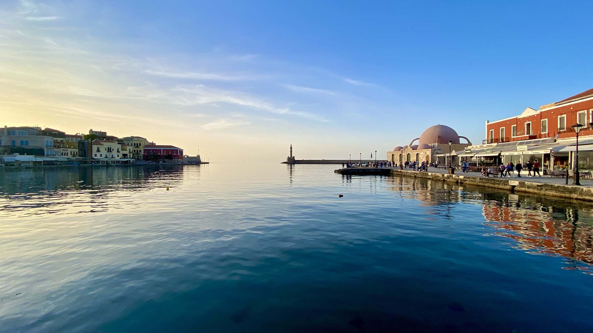 
Calm waterfront view of Chania's Venetian Harbor at sunrise, with lighthouse, domes and waterfront cafes 