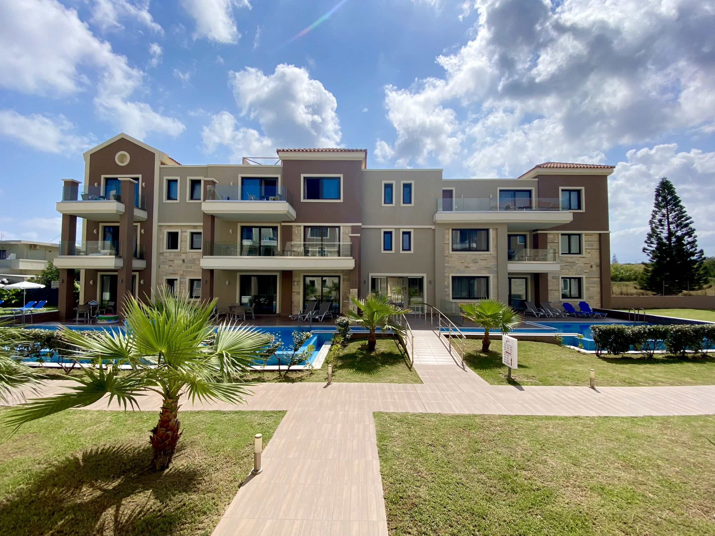 
Modern three-story apartment building with balconies, pools and palm trees under a bright sky