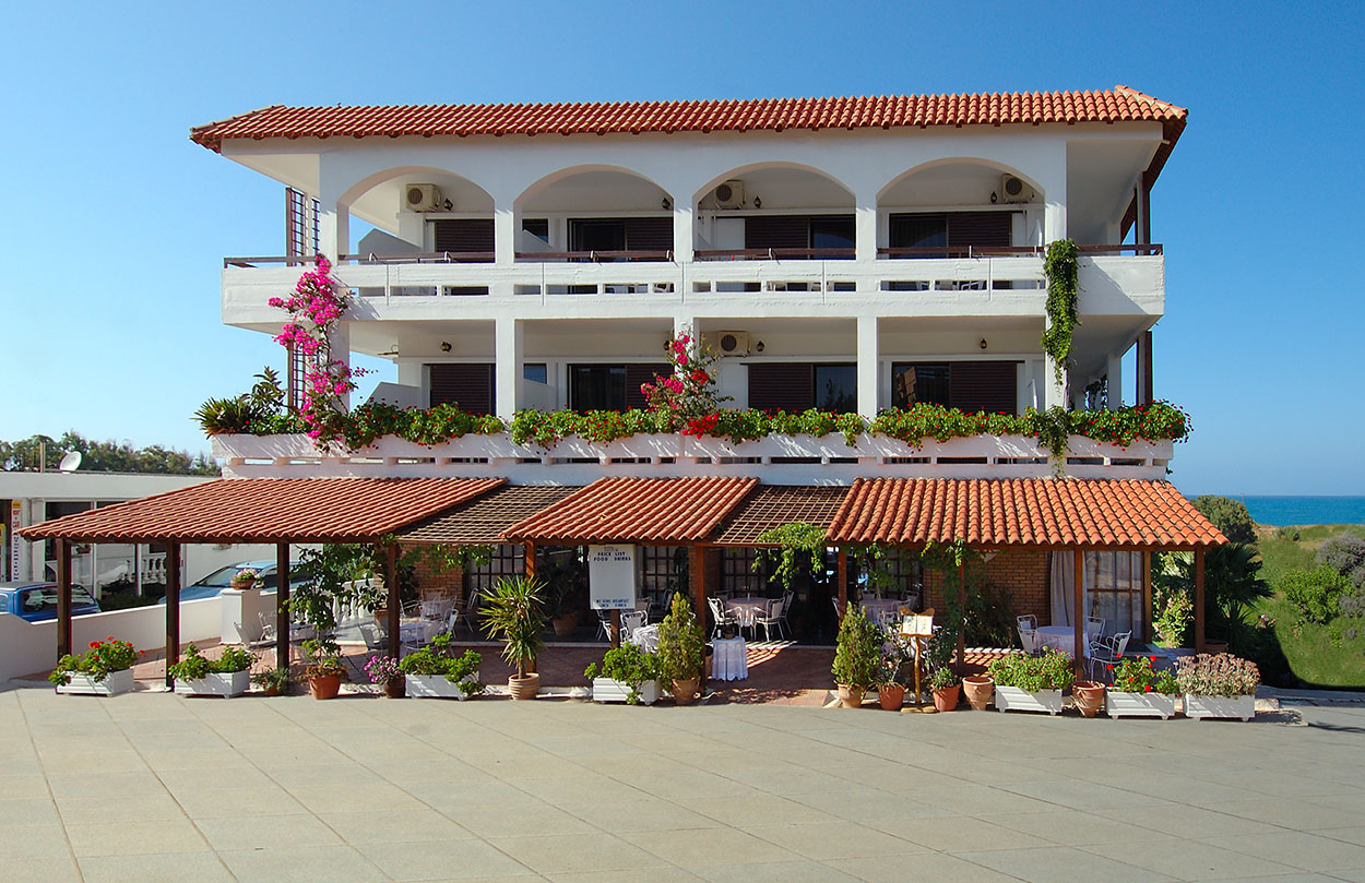 
White three-story hotel building with terracotta roof, balconies and vibrant flowers overlooking the sea