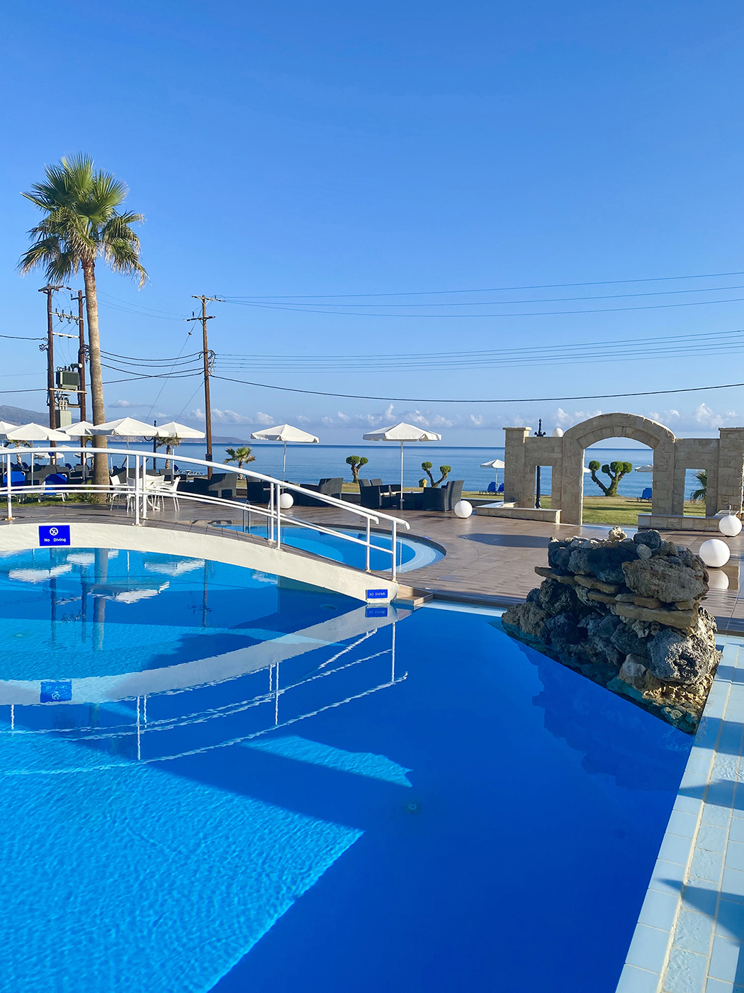 
Bright blue swimming pool with a small bridge, sea views, and white umbrellas under a clear sunny sky