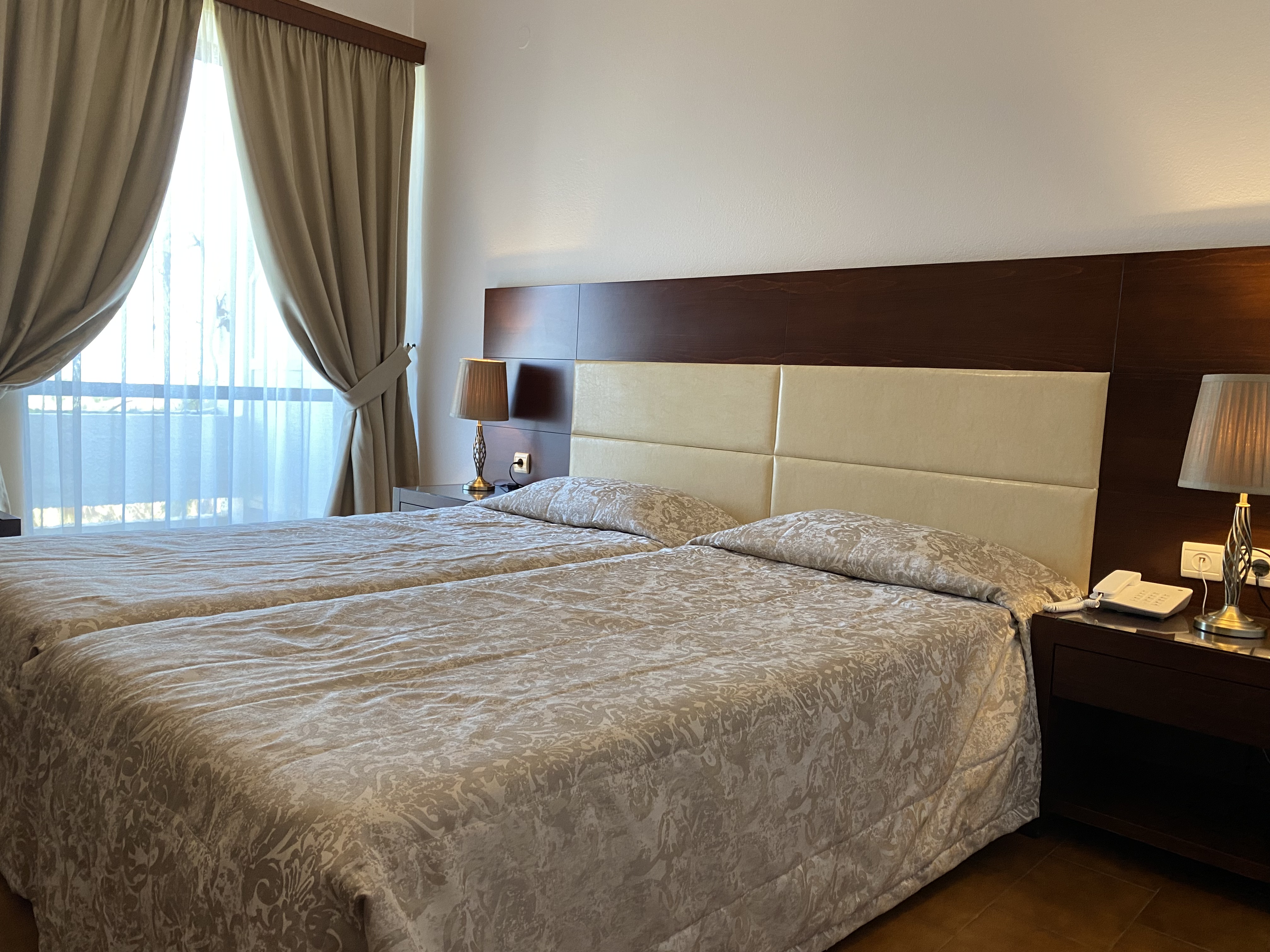 
A comfortable double room with two neatly made beds, elegant bedside lamps, and large curtains letting in natural light