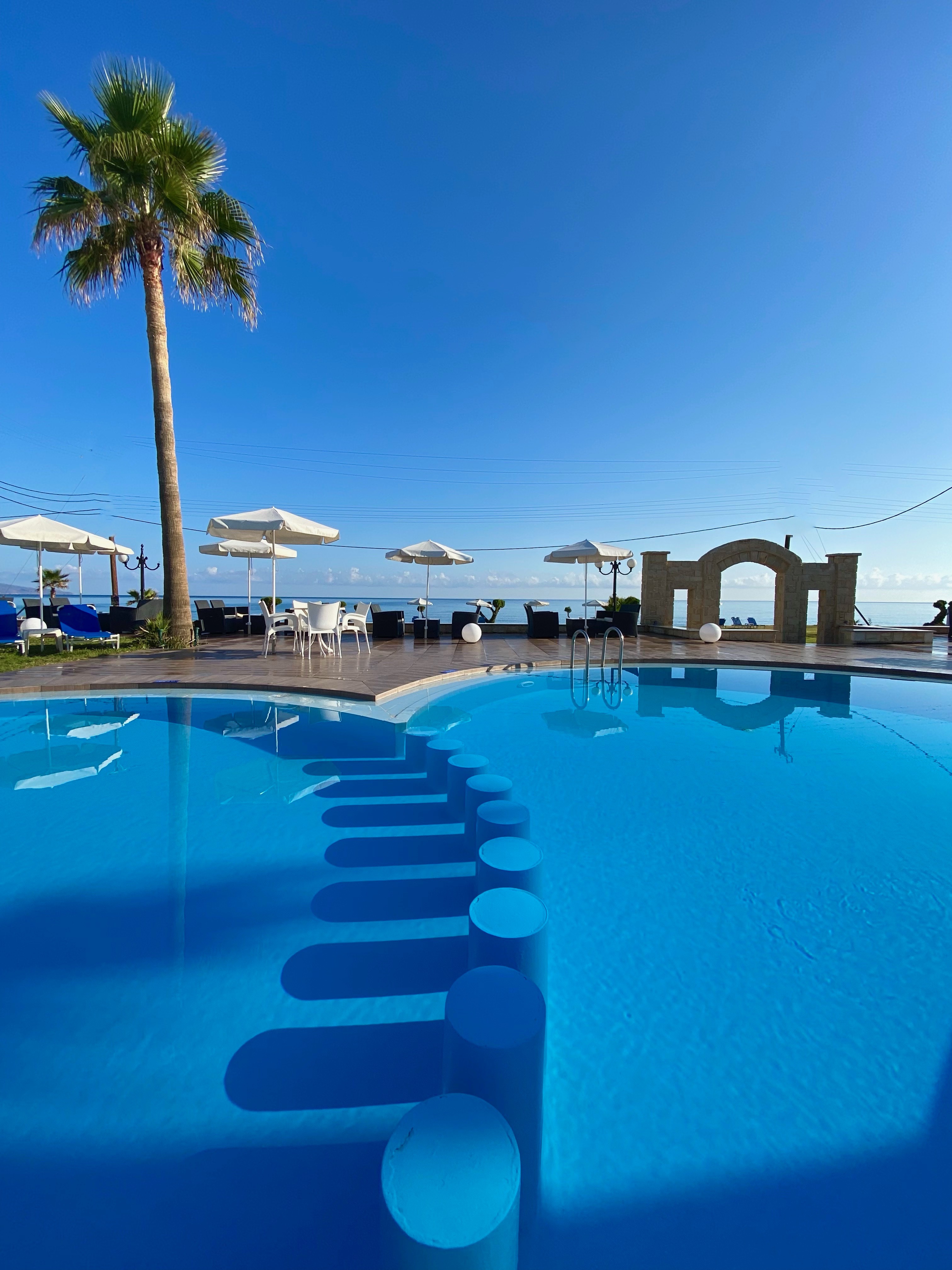 
Main pool with built-in seating, surrounding tables with umbrellas, a palm tree and sea view in the background