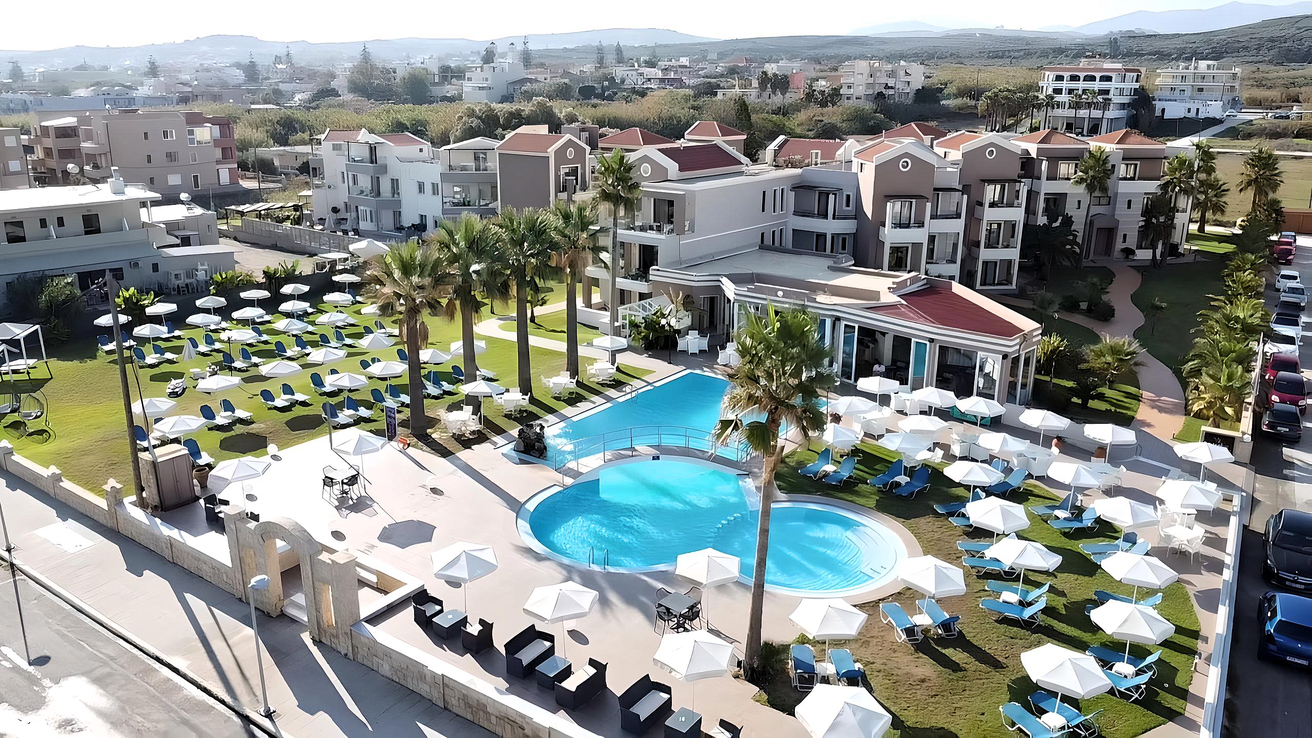 
Aerial view of Mike apartment complex with lawn with sunbeds, a central pool and palm tree garden